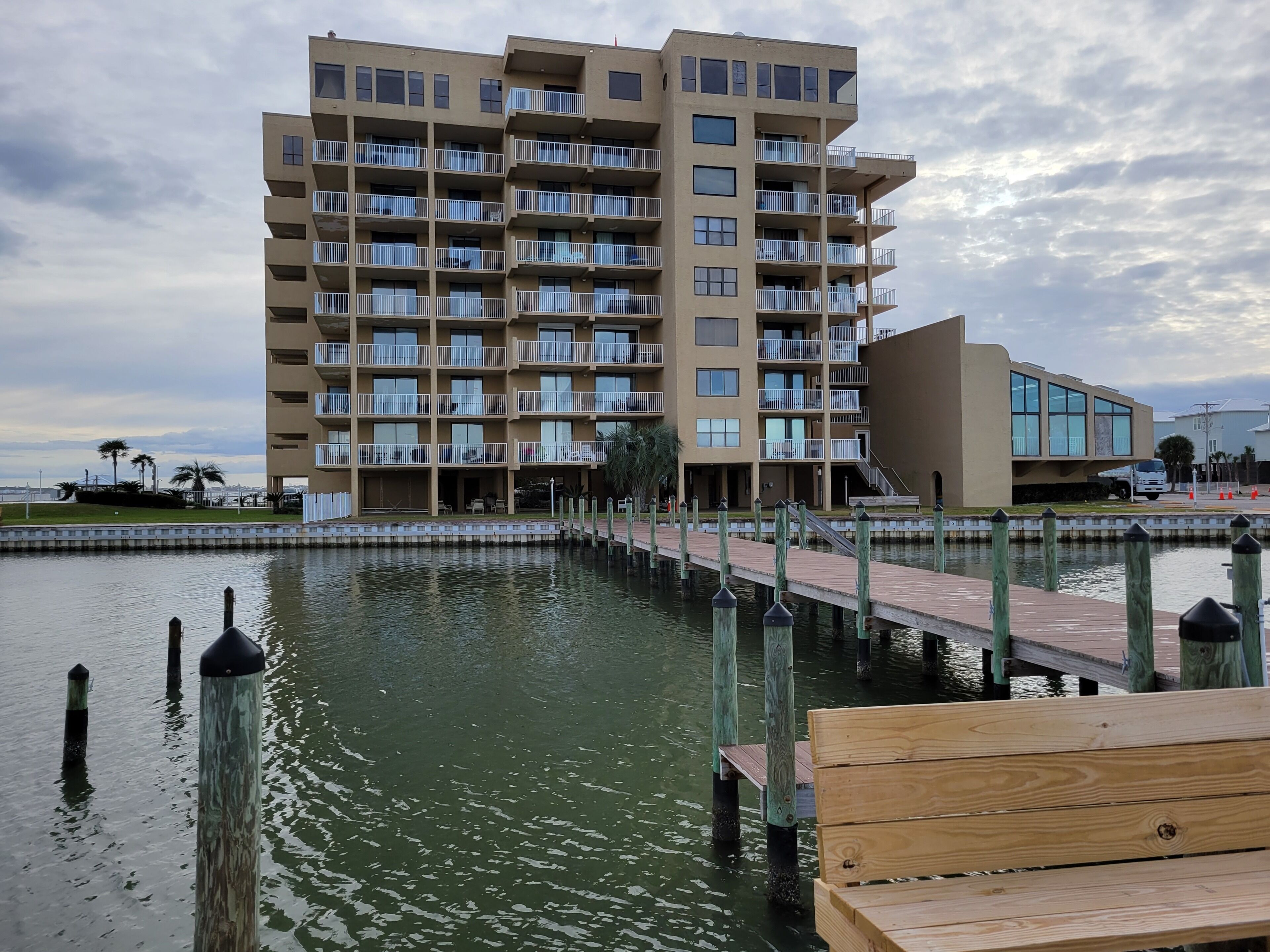 Un immeuble à plusieurs étages doté de balcons, une jetée s’avançant dans une étendue d’eau et une petite construction en bois sur la jetée.