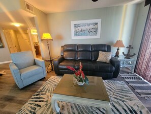 A living room with a black leather sofa, a light blue armchair, a coffee table with a vase of flowers, and a framed picture on the wall.