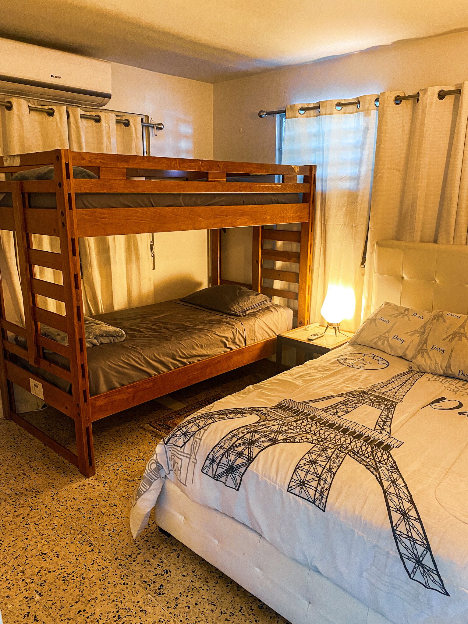 A room with a bunk bed, a single bed with Eiffel Tower-themed bedding, a bedside lamp, and a window with curtains.