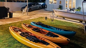 Una casa de dos pisos con un patio techado, un barco sobre un remolque y kayaks en el césped.