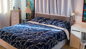 Un dormitorio con cama, ventilador de techo, mesita de noche, lámpara, un cuadro y una ventana con cortinas.