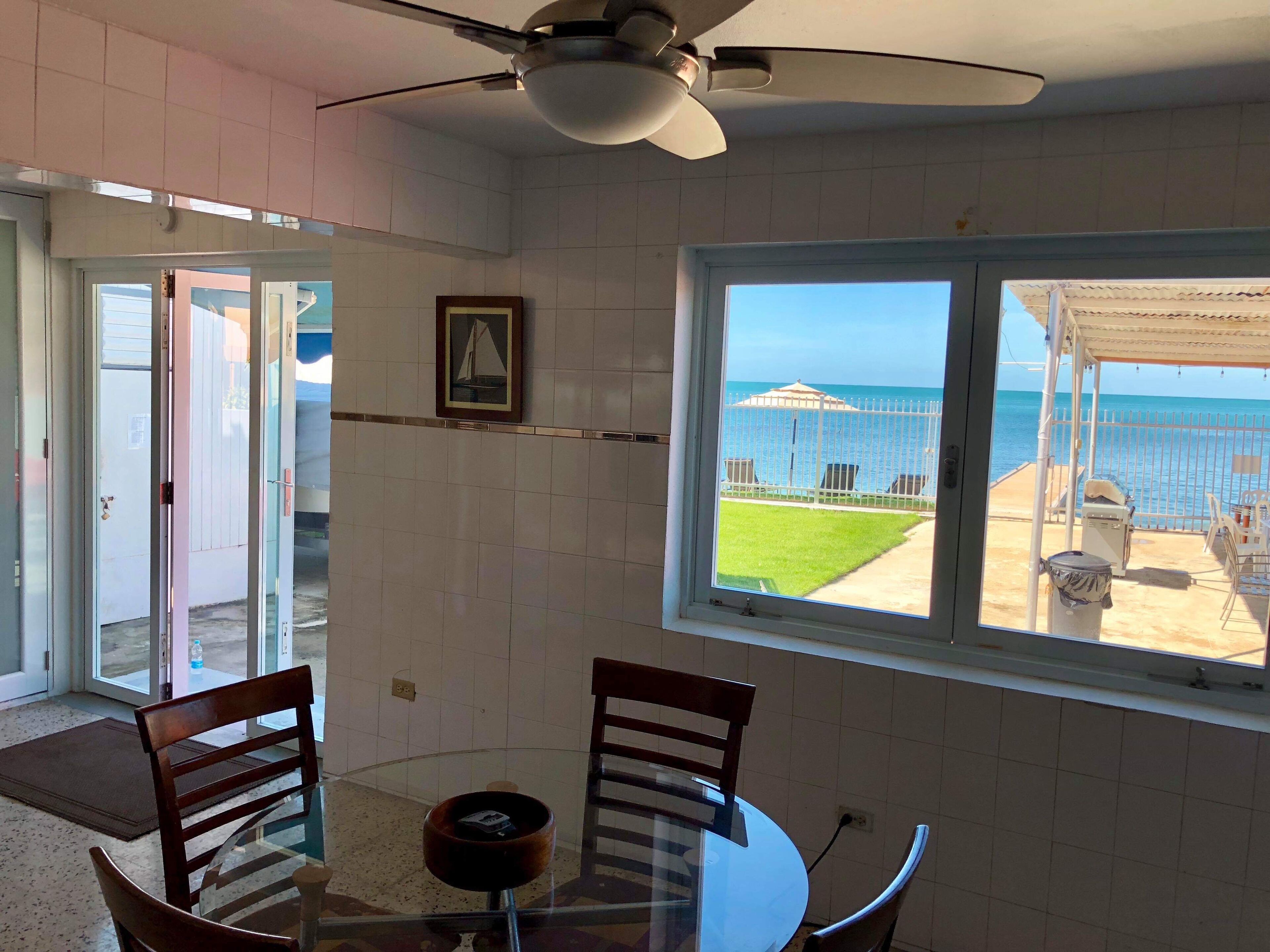 A room with a glass table, two wooden chairs, a ceiling fan, and a window with a view of the sea and a beach.