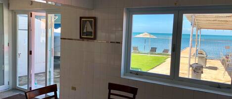 A room with a glass table, two wooden chairs, a ceiling fan, and a window with a view of the sea and a beach.