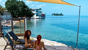 Un espacio de descanso junto a la playa con sillones y una zona sombreada, con vistas a un mar tranquilo y una isla distante.