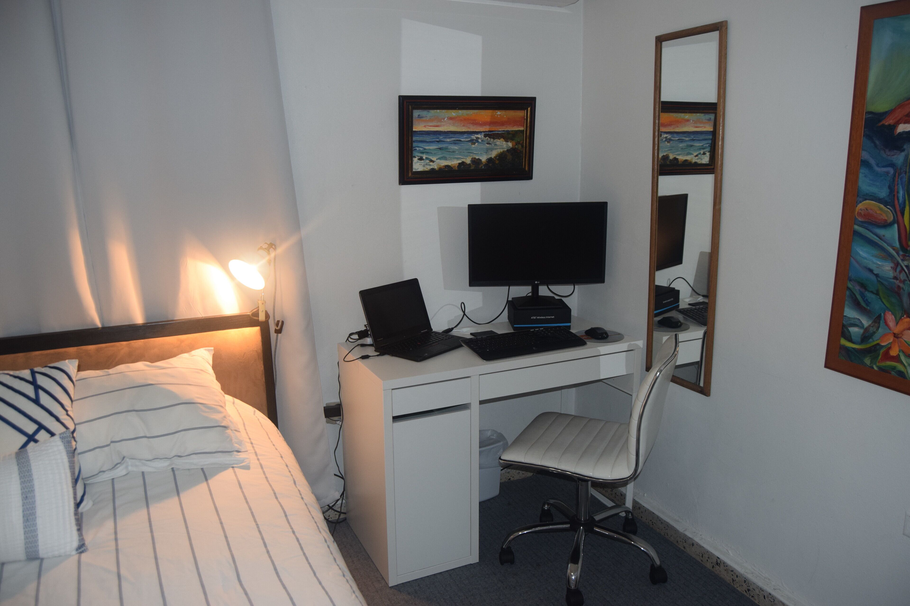 Un dormitorio con una cama, un escritorio con una computadora portátil, una silla y dos cuadros en la pared.