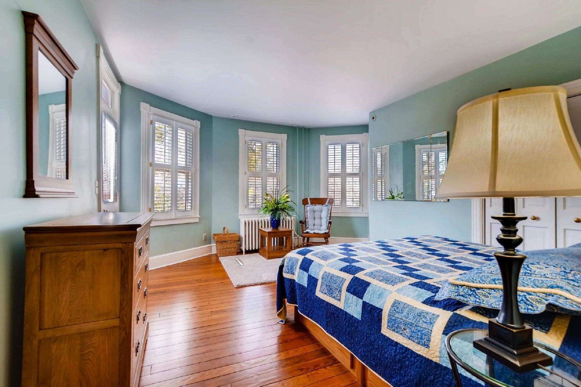 Un dormitorio con un aparador de madera, una cama con colcha azul, una silla, una mesa con una planta y una ventana con contraventanas.
