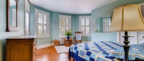 Un dormitorio con un aparador de madera, una cama con colcha azul, una silla, una mesa con una planta y una ventana con contraventanas.