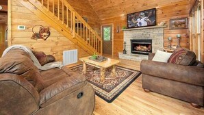 A log cabin interior with a wooden staircase, a fireplace, a television mounted above it, and two brown leather sofas. - Moondance: Pet Friendly - Brand New Hot Tub - Cozy Wooded Atmosphere (Sevierville)