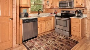 A wooden kitchen with a dishwasher, stove, microwave, and a window with blinds. - Moondance: Pet Friendly - Brand New Hot Tub - Cozy Wooded Atmosphere (Sevierville)