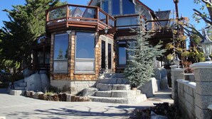 A modern house with a large glass bay window, stone exterior, and a small evergreen tree in front.