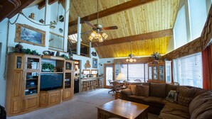 A spacious living area with a high ceiling, wooden beams, and a ceiling fan. There is a dining area with a table and chairs, and a kitchen area with cabinets and a television.