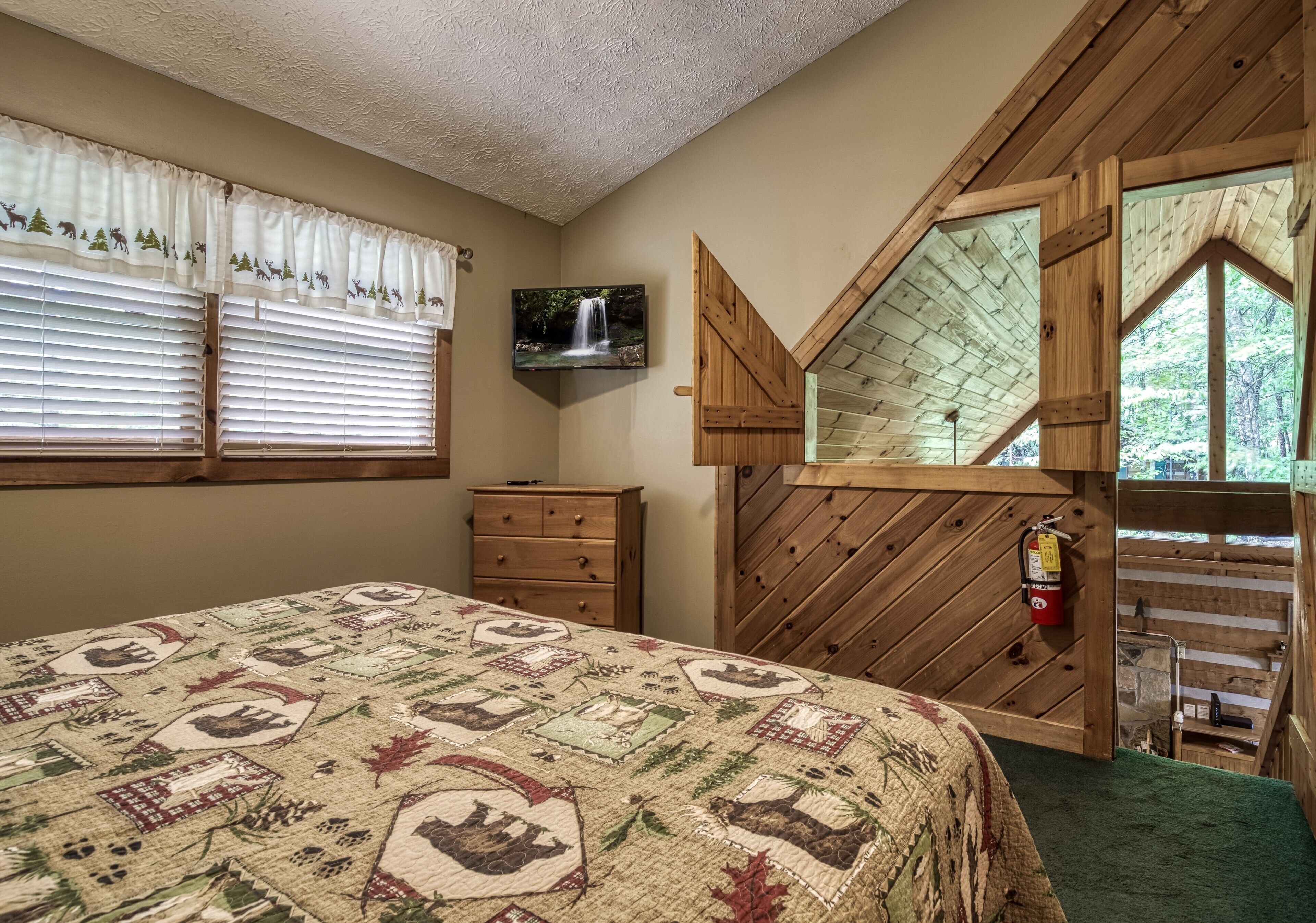 A bedroom with a bed, a dresser, a television, a window with a view of a waterfall, and a wooden loft area.