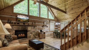 A cozy cabin interior with a stone fireplace, wooden beams, a large window, and a staircase.