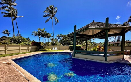 A swimming pool with clear blue water, surrounded by palm trees and a covered pavilion.