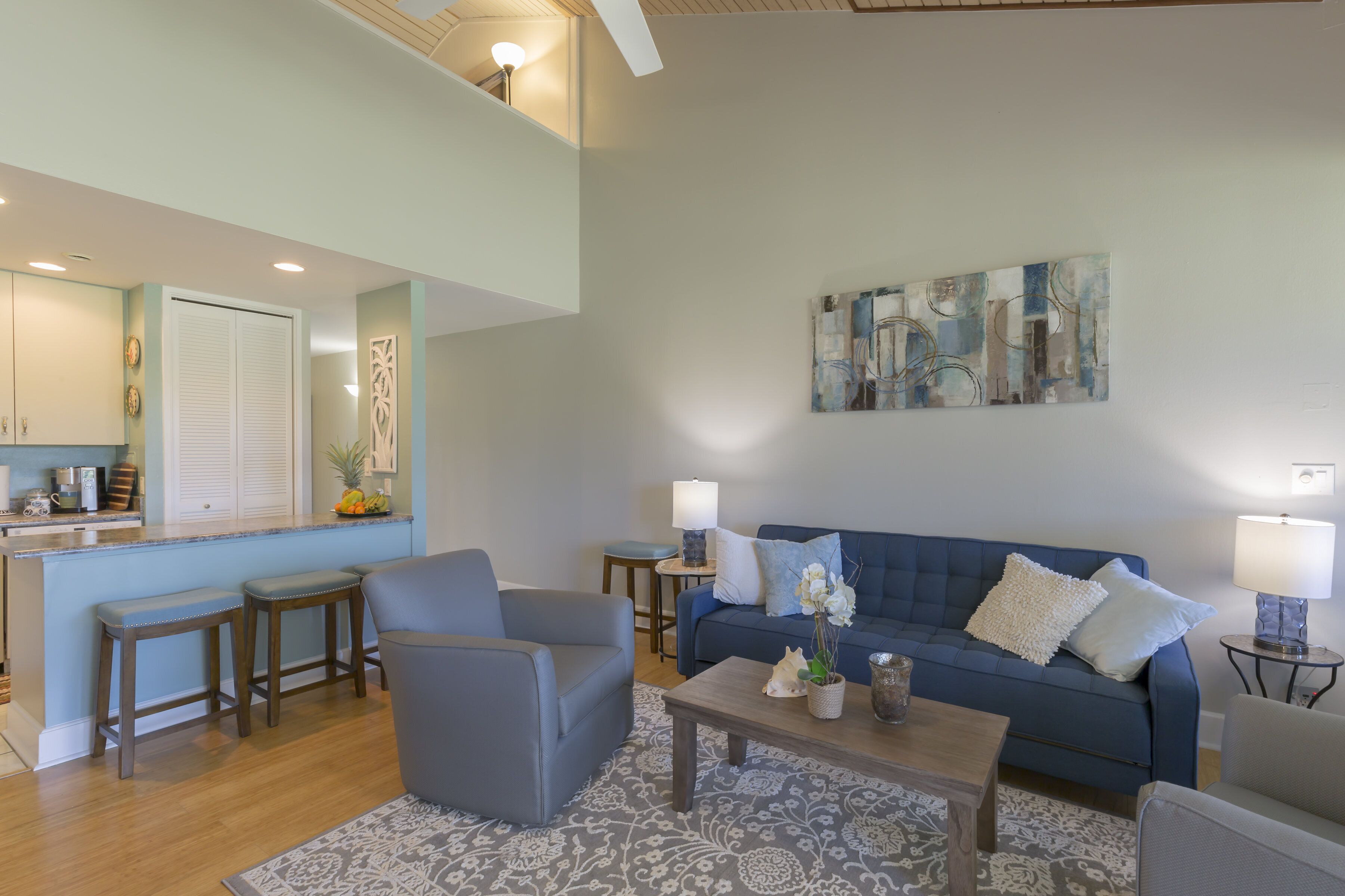 Una sala de estar moderna con un sofá azul, dos sillones, una mesa de centro y un área de cocina con mostrador y taburetes.