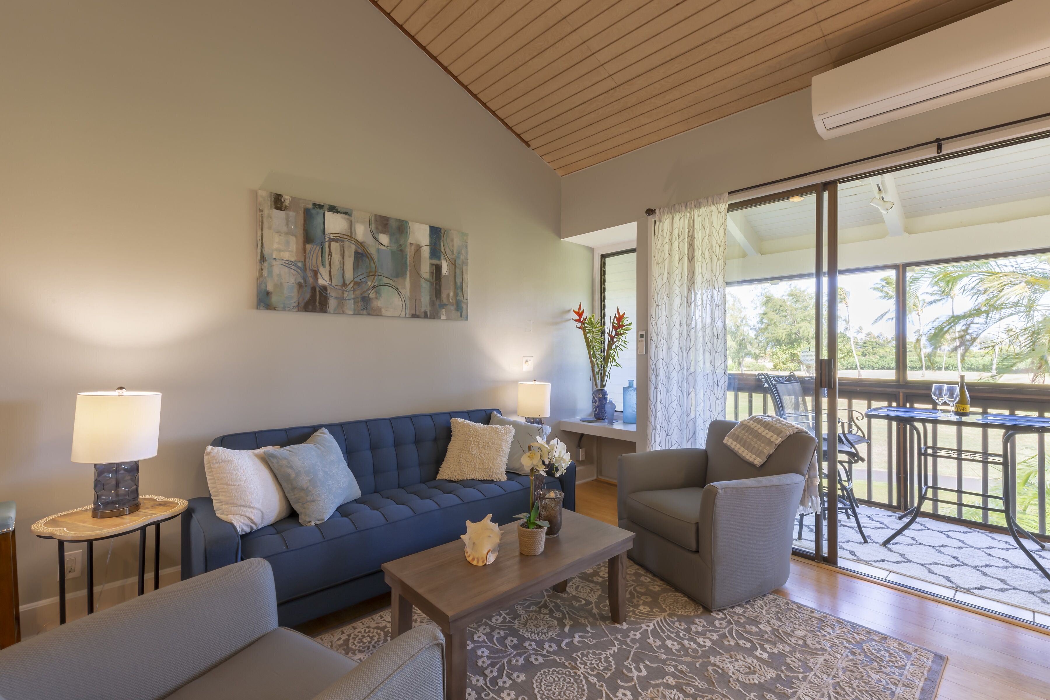 Una sala de estar moderna con un sofá azul, un sillón gris, una mesa de centro de madera y un tapete estampado. En la pared hay un cuadro, una lámpara sobre una mesita auxiliar y vistas al exterior a través de amplios ventanales.