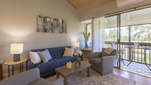 A modern living room with a blue sofa, a grey armchair, a wooden coffee table, and a patterned rug. There is a painting on the wall, a lamp on a side table, and a view of the outdoors through large windows. - Resort Living Off the Beaten Path
AC , WIFI, and Fully Remodeled Bathroom (Kahuku)