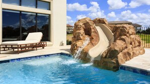 A swimming pool with a water slide, lounge chairs, and a view of a house in the background. - Waterfront Home-Bowling Alley, Cinema, Sauna, Outdoor Grilling, Pool w/ Slide  (Four Corners)