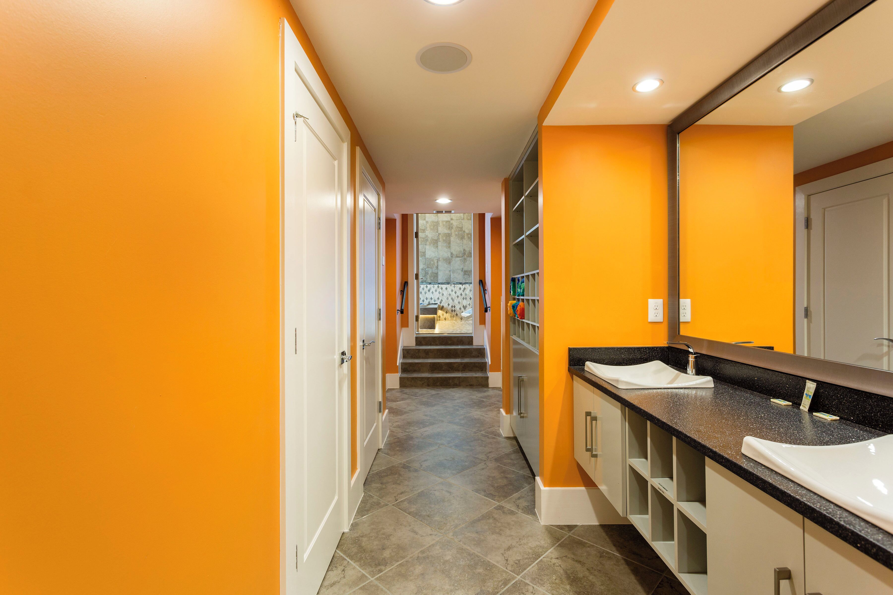 Un pasillo con un baño que incluye un lavabo y un espejo, suelo de baldosas y una escalera que lleva a otra habitación.
