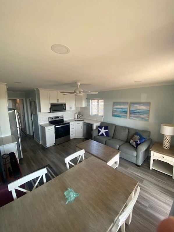 A modern kitchen with white cabinets, a microwave, and a stove. There is a dining area with a wooden table and chairs. A gray sofa and a small white table with a lamp are in the living area.