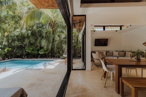A modern interior with a pool, a dining area, and a living space with a view of tropical vegetation.