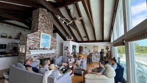 A spacious living room with a fireplace, wooden beams, and large windows overlooking a deck.