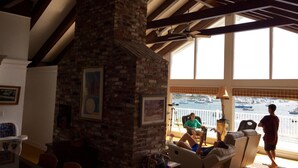 A cozy room with a brick fireplace, wooden beams, and a view of boats through large windows.