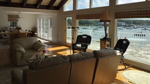 A spacious living room with a large window overlooking a marina. There is a sofa, two chairs, and a dining area with a table and chairs.