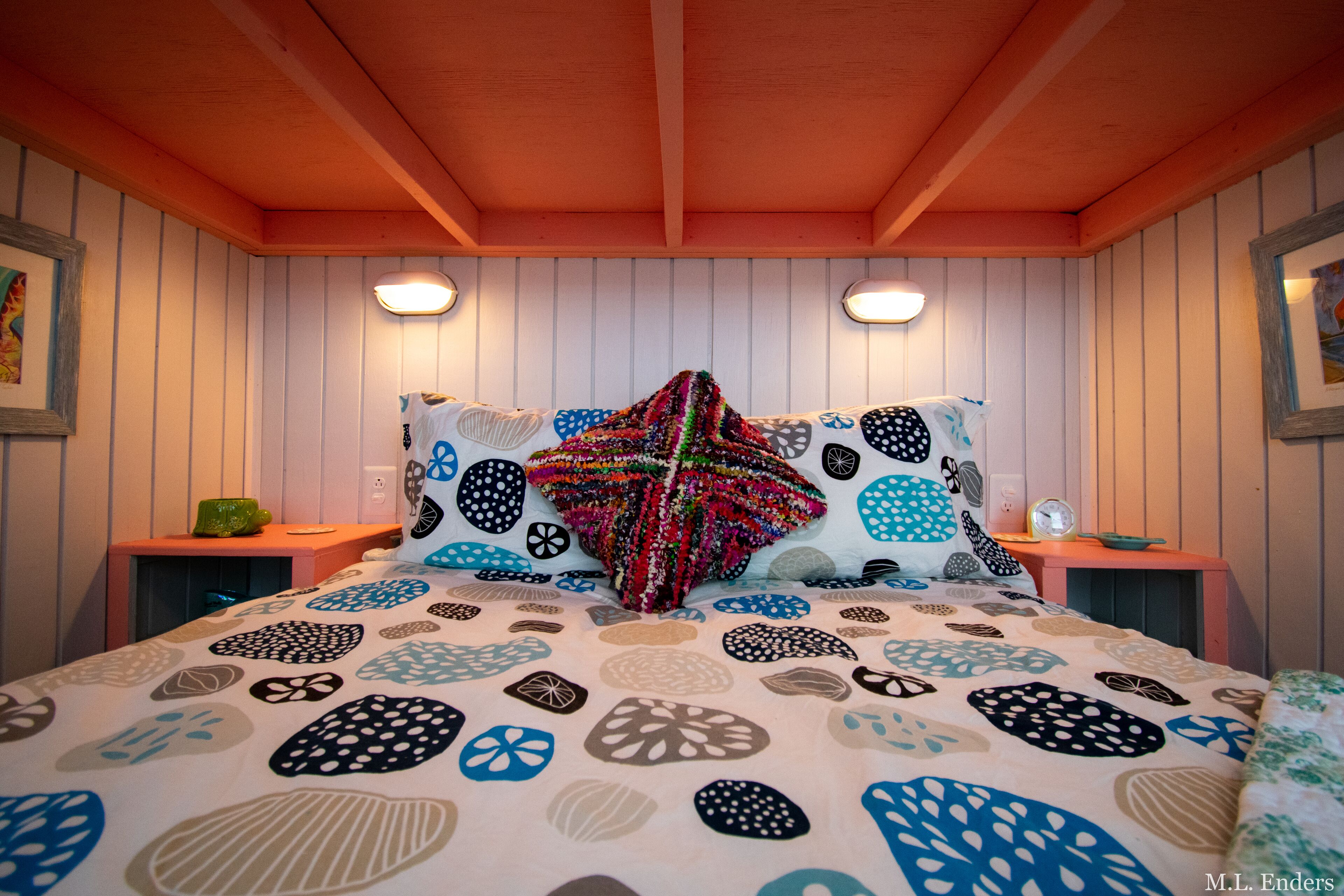 Ein Schlafzimmer mit einem Bett, das mit einer gemusterten Decke bezogen ist, zwei Nachttischen mit Lampen und einer Holzbodenverkleidung.