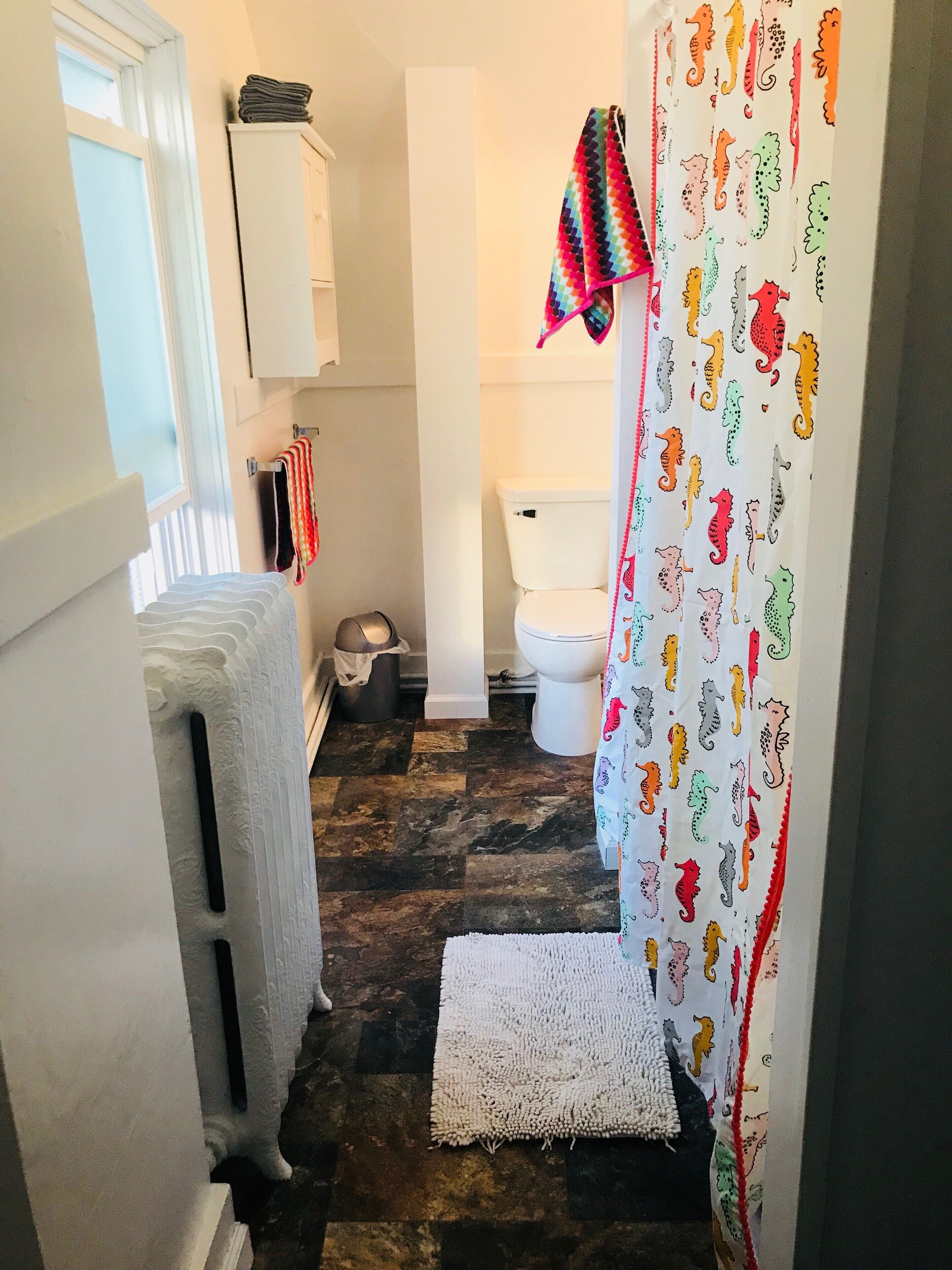 Un baño con inodoro blanco, radiador blanco, alfombra blanca y cortina de ducha con motivos de caballitos de mar.