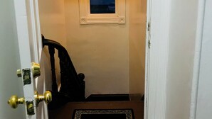 Un couloir étroit avec une porte sur la gauche, un paillasson noir sur lequel est écrit « Bienvenue », une fenêtre donnant sur le ciel et un luminaire suspendu.