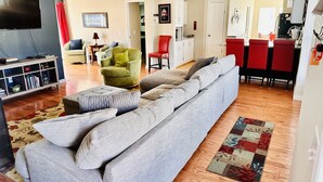 Una sala de estar con un sofá gris, un sillón verde y un televisor de pantalla plana. Se observa un comedor con sillas rojas y una cocina al fondo.