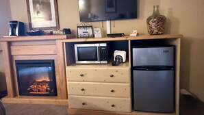 A living room with a fireplace, a flat-screen TV mounted above it, a microwave, a refrigerator, and a wooden cabinet with drawers.