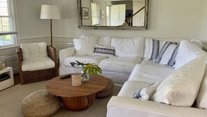 A living room with a white sectional sofa, a wicker chair, a round wooden coffee table, and a large mirror on the wall. - Family Friendly Island Getaway (Nantucket)