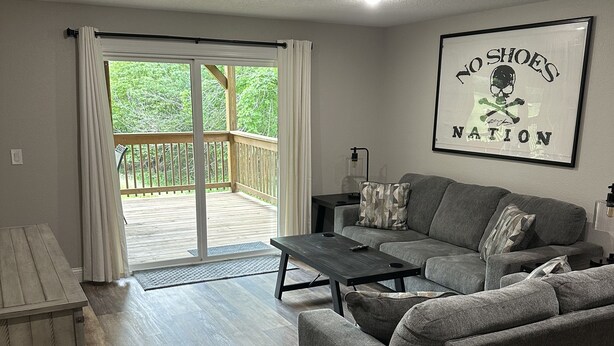 A living room with a grey sofa, a coffee table, and a wooden deck with a glass door leading outside. - Branson area Lakehouse, 5 Bedrooms, 3 Baths, (Sleeps 9-15) New decks Spring 2019 (Kimberling City)