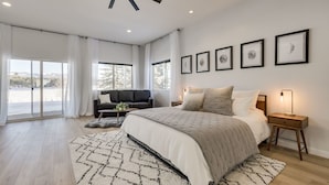 A modern bedroom with a large bed, a sofa, and a desk. There are windows with curtains, a ceiling fan, and framed pictures on the wall.
