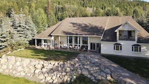 A large, two-story house with a stone retaining wall in front, surrounded by dense forest.