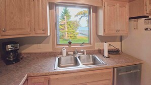 Una cocina con armarios de madera, un lavamanos doble, una ventana con vista a árboles y un cuerpo de agua, y una cafetera sobre la encimera.