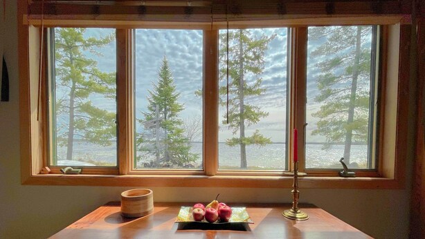 Una mesa de madera con una vela, un libro y manzanas junto a una ventana que da a árboles y agua.