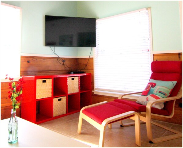 A room with a television mounted on the wall, a red storage unit with wicker baskets, a red cushioned chair with a cushion featuring a vintage car design, and a small table with a clear glass bottle. - Walk to the beach! Heart of Ocean Beach! (San Diego)