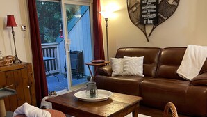 Een gezellige woonkamer met een bruine leren bank, een houten salontafel en een rieten fauteuil. Een poef met rood-wit patroon staat op de voorgrond.