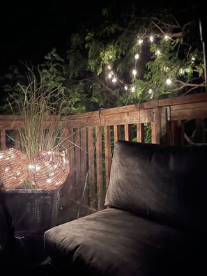 Eine Holzterrasse mit gepolsterten Sitzgelegenheiten, umgeben von Grünpflanzen und Lichterketten.