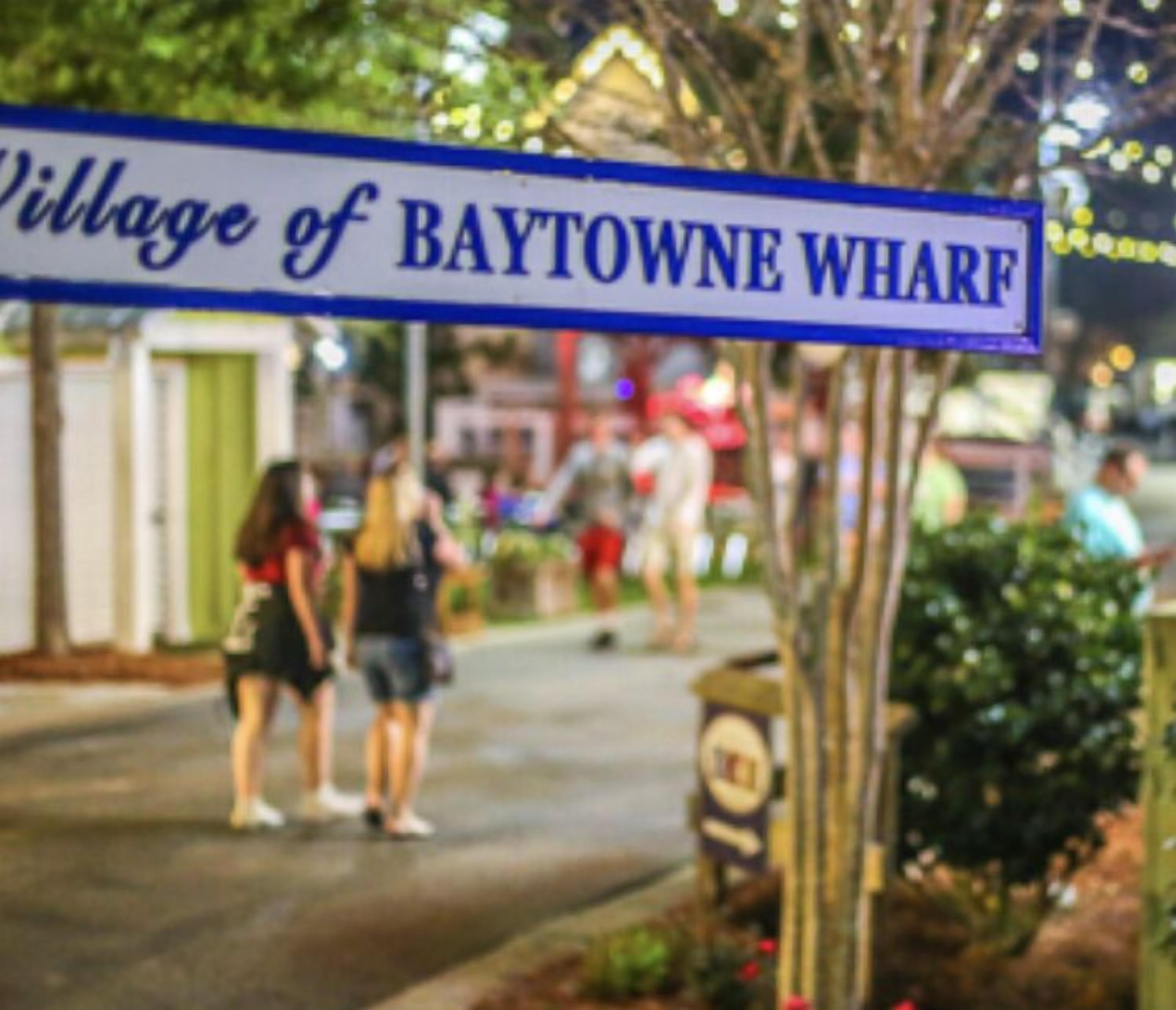 A street sign reading "Village of Baytowne Wharf" with a blurred background of people and greenery.