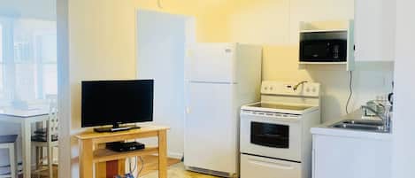 Ett kompakt bostadsutrymme med en TV på ett träställ, ett vitt kylskåp, en mikrovågsugn, en spis och en liten köksbänk.