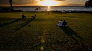 Un campo cubierto de césped con gente sentada y caminando, un atardecer en el fondo y una masa de agua.