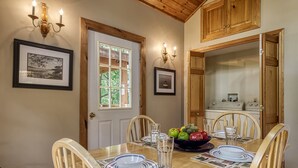 A dining area with a wooden table set for four, surrounded by chairs. There are framed pictures on the walls, a window with a view of greenery, and a kitchen area visible through an open door. - Lake Front with Lake view, King size Bed Great Place to relax and go fishing (Sevierville)