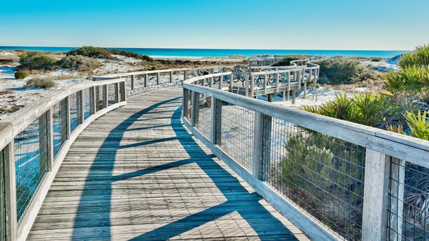 Une promenade en bois dotée d’une rambarde métallique, qui mène à une plage aux eaux d’un bleu limpide.