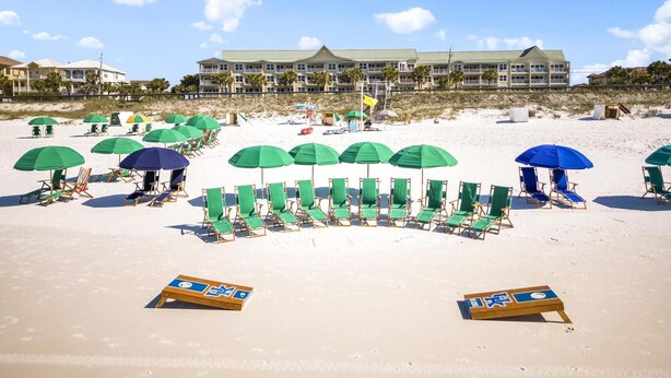 Una spiaggia con file di sedie a sdraio e ombrelloni verdi e blu, un grande edificio di un hotel sullo sfondo e un'ampia area di spiaggia sabbiosa.