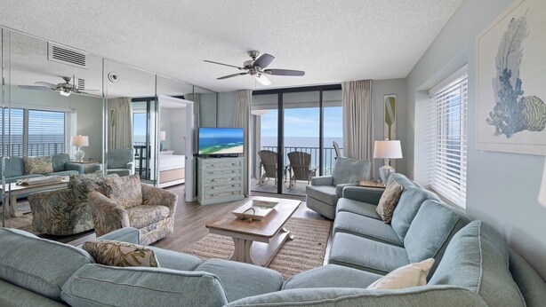 A spacious living room with a large flat-screen TV, comfortable seating, a ceiling fan, and a view of the ocean through large windows.