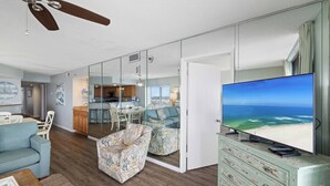 A modern living room with a large flat-screen TV, a ceiling fan, and a view of the ocean through a glass door.